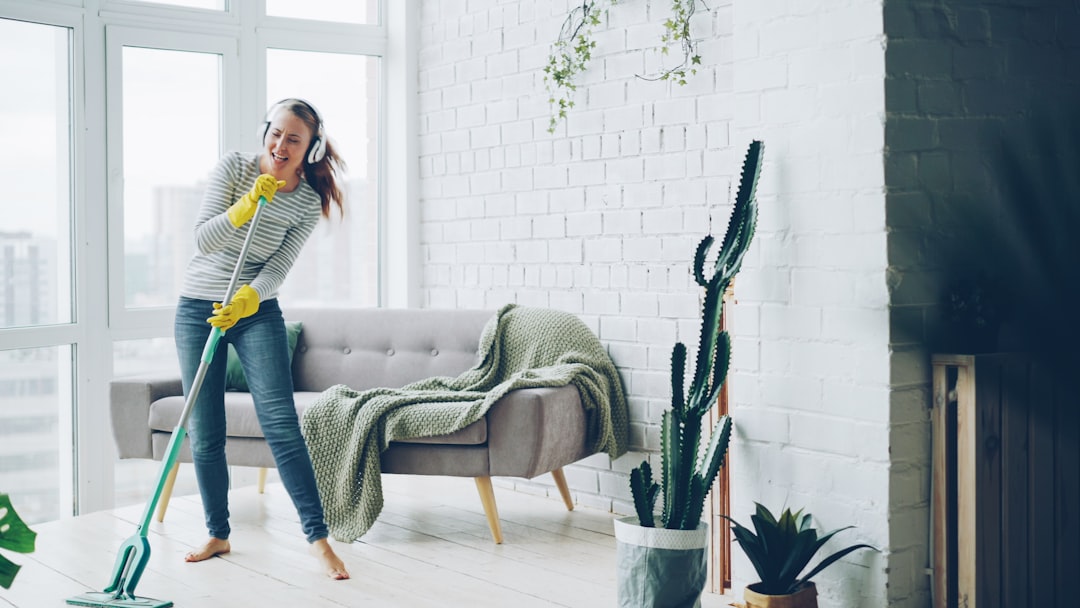 Pretty blond girl is washing floor with flat plastic mop and listening to music in headphones, dancing and singing during routine housework in beautiful apartment.